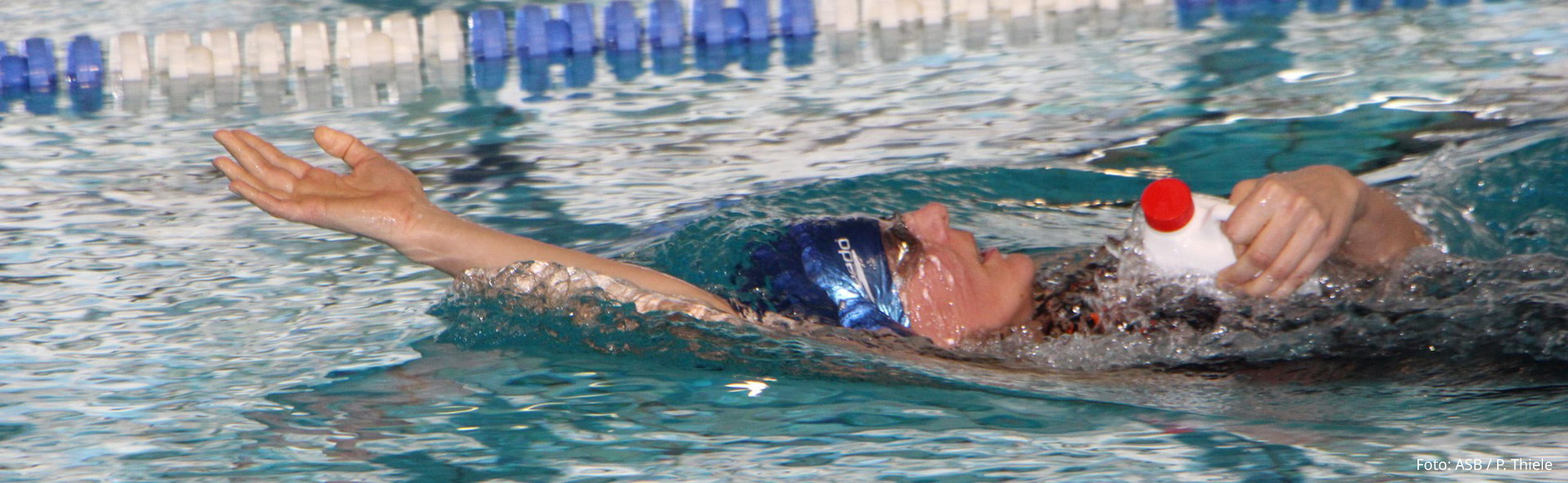 Schwimmerin schwimmt einarmig auf dem Rücken mit Flasche in der Hand in Schwimmhalle